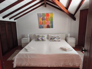 A bedroom with a sloped ceiling, a bed with a white bedspread, two decorative pillows, a colorful abstract painting on the wall, and wooden wardrobes. - 2-bedr. Harmony villa with garden 250 meters from Patacho sea beach (Vila Nova de Milfontes)