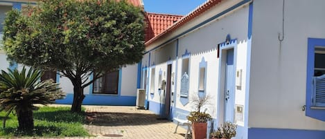 A courtyard with a tree, potted plants, a bench, and a building with blue and white walls and a red-tiled roof.