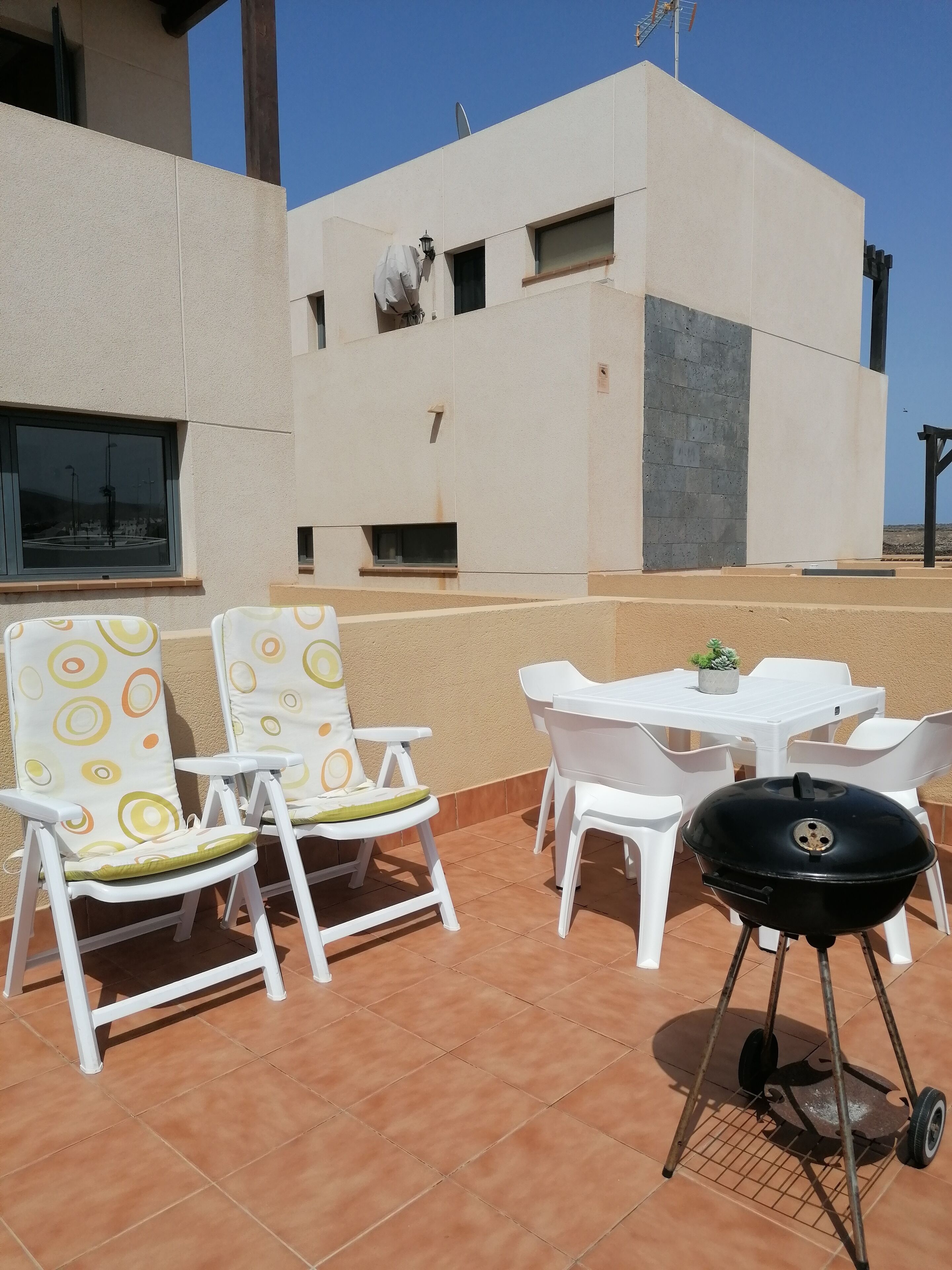 Un patio al aire libre con sillas de plástico blanco y una mesa, una parrilla negra y una planta en maceta.
