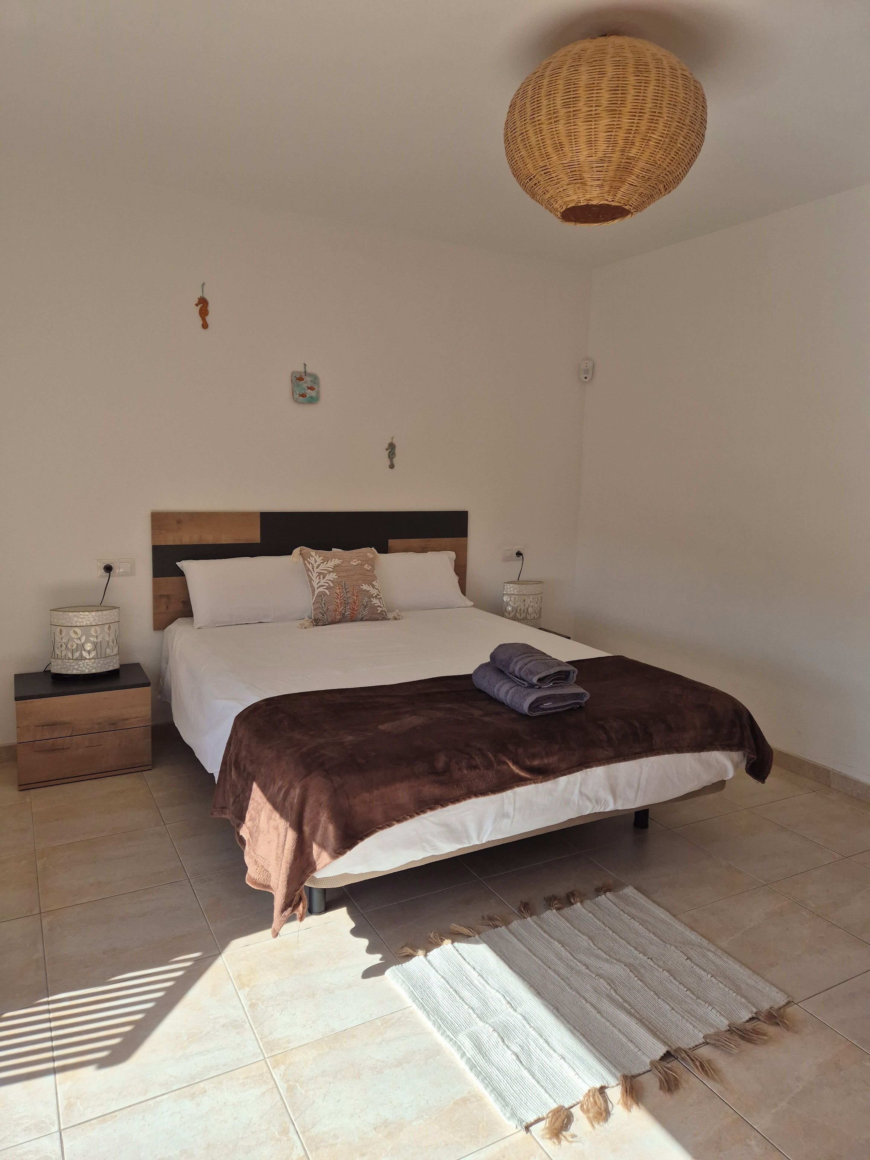 A neatly made bed with a brown blanket, a bedside lamp, and a woven light fixture above it.