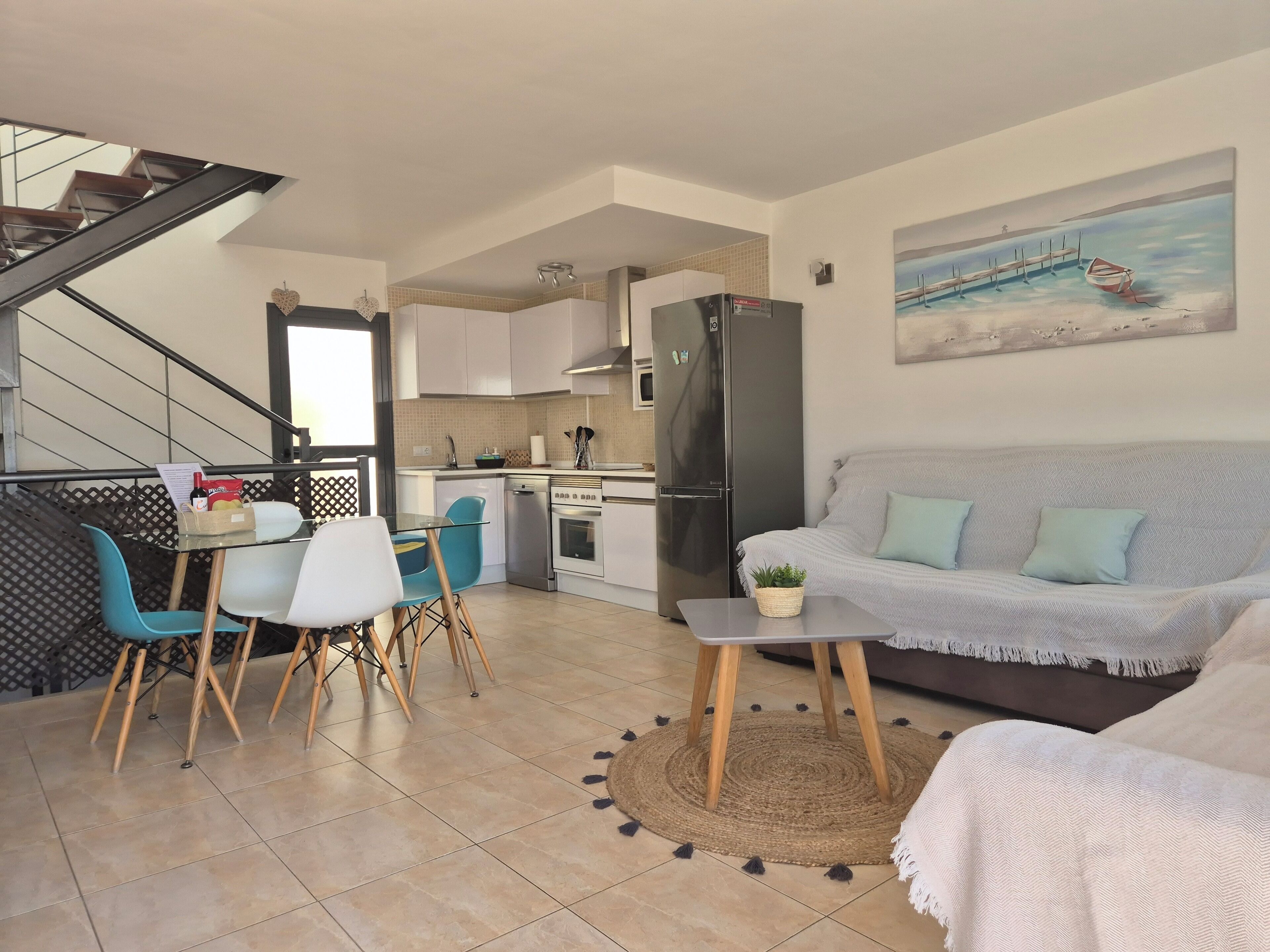 A modern kitchen with a refrigerator, stove, and dishwasher. There is a dining area with a glass table and chairs. A living room with a sofa and a painting on the wall.