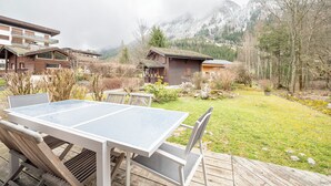 Une terrasse en bois avec une table et des chaises à plateau de verre, offrant une vue sur un paysage montagneux avec des bâtiments en arrière-plan.