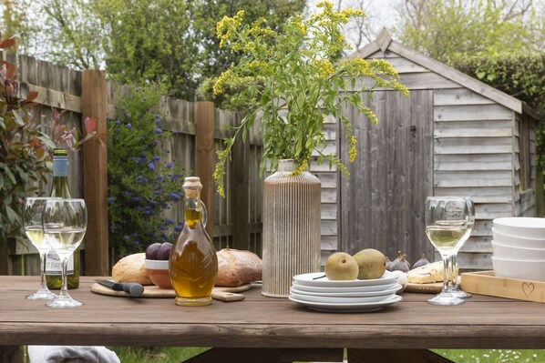 A wooden table with a bottle of olive oil, a loaf of bread, a bowl of grapes, a vase with yellow flowers, and plates with cheese and pears. - A stunning holiday home located in the Curved Stables at Roserrow. (Wadebridge)