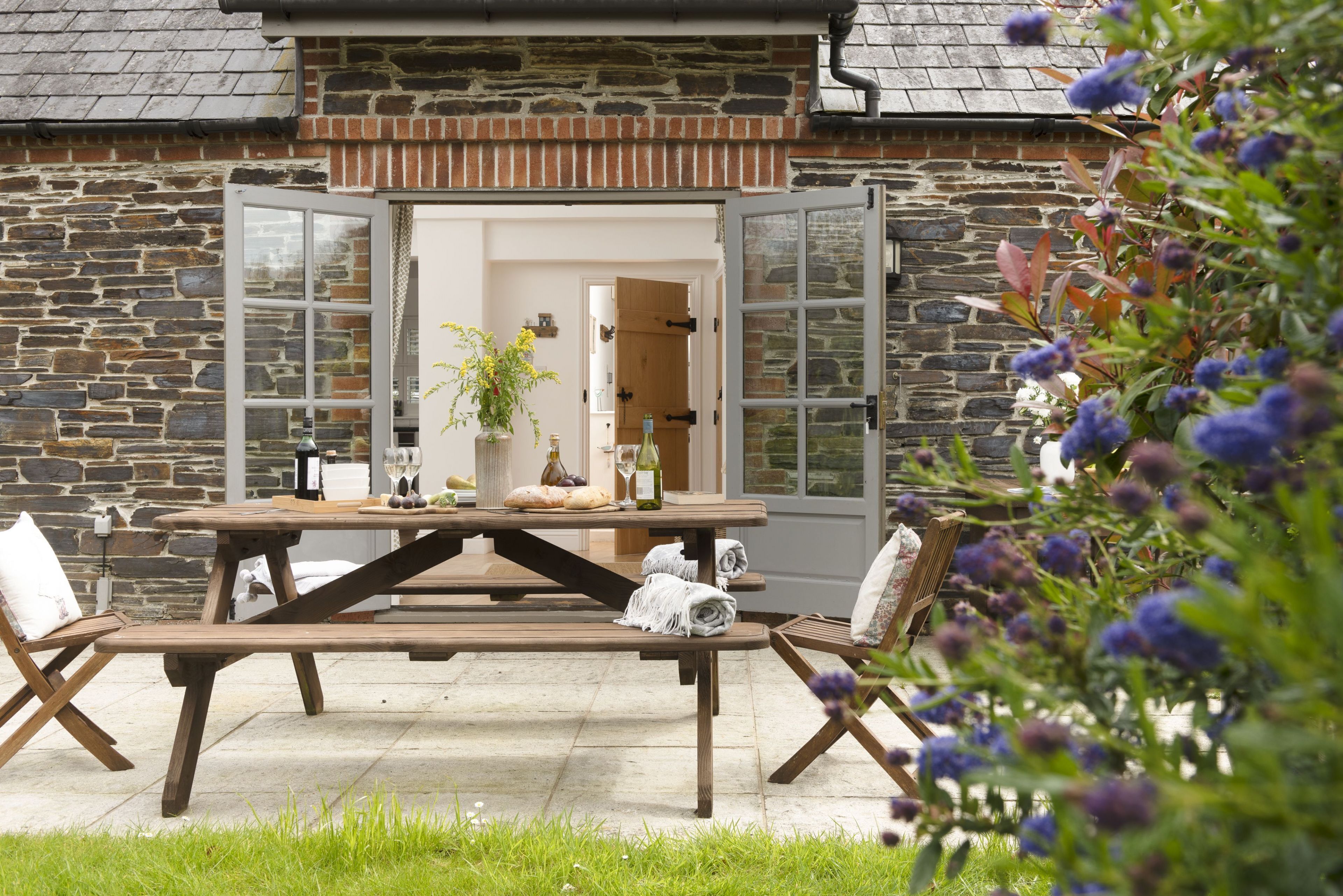 An outdoor dining area with a stone wall, wooden picnic table, and chairs.