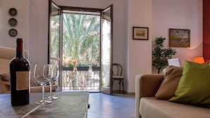 Un salon avec un canapé, une table sur laquelle se trouvent une bouteille et des verres, un fauteuil, une plante et une vue sur des palmiers, visible à travers une porte vitrée.