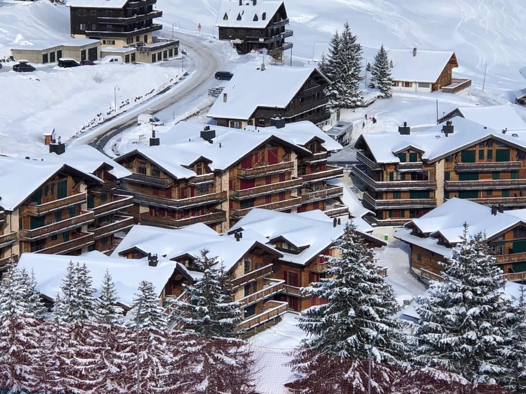 Een besneeuwde vakantiebestemming met meerdere chalets, een kronkelende weg en sneeuwrijke bomen.