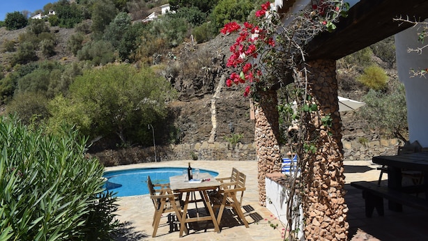 A pool area with a stone patio, wooden table and chairs, and a stone pillar with climbing plants. - Finca Carrulo with private pool for 6 people and fantastic views (Cómpeta)