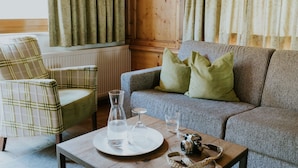 Una sala de estar acogedora con un sillón escocés, un sofá gris con cojines verdes y una mesa de centro de madera con un vaso de agua, una copa de vino y una cámara.