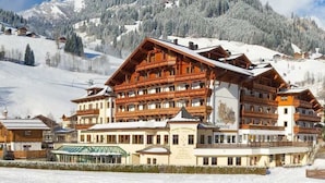 Un hotel grande de varios pisos con arquitectura alpina tradicional, que cuenta con balcones y fachadas de madera, frente a un paisaje montañoso nevado.