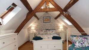 Une chambre avec un plafond en pente, des poutres en bois, un mur en briques, un lit avec une literie à motifs floraux, une commode et une table de chevet avec une lampe.