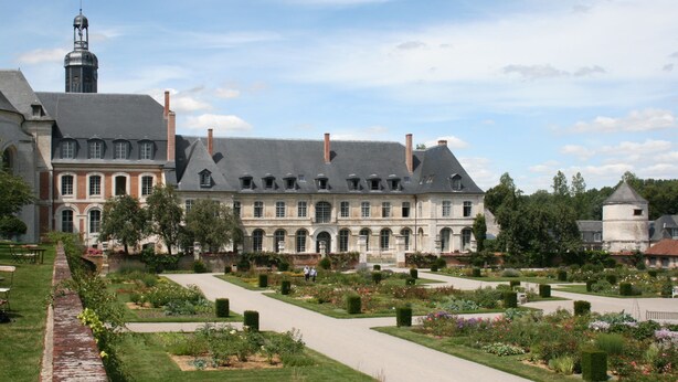 Un grand bâtiment historique doté d’un jardin à la disposition symétrique, comprenant un chemin central et divers parterres de fleurs.