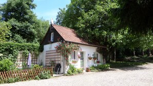 Un petit bâtiment blanc, surmonté d’un toit recouvert de tuiles rouges, entouré d’une végétation luxuriante et bordé d’une allée en gravier.