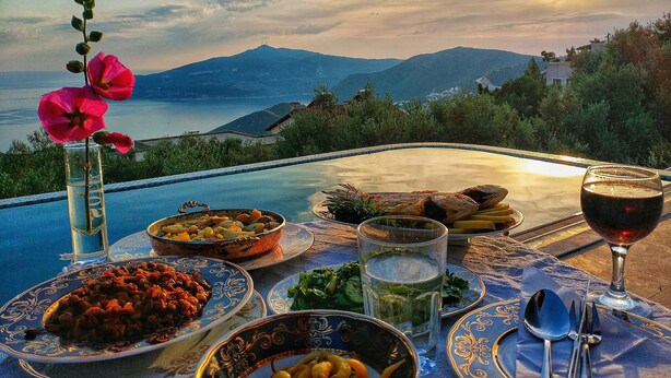 Une table à manger dressée avec des assiettes de nourriture, un verre de vin et un vase rempli de fleurs, offrant une vue panoramique sur des montagnes et un plan d’eau.