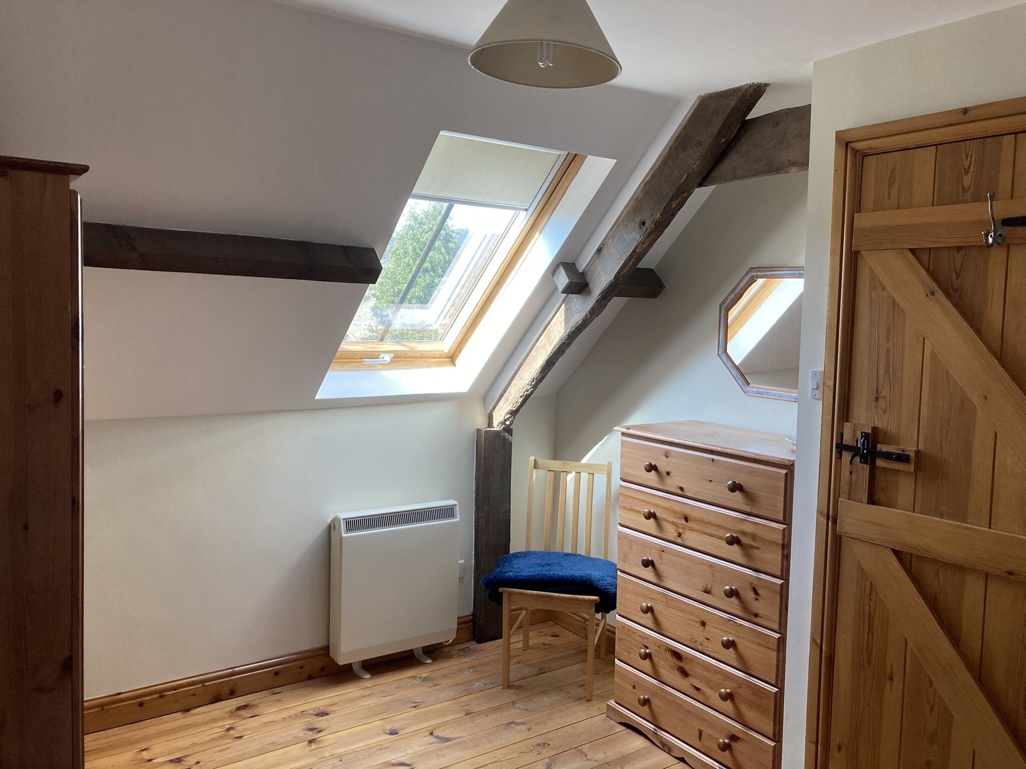 Ein Zimmer mit schrägen Wänden, Holzbalken, einem Oberlicht, einer Heizung, einem hölzernen Schminktisch und einem Stuhl.