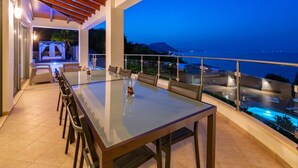 Un espace repas avec une table et des chaises en verre, donnant sur une piscine et un paysage montagneux au crépuscule.