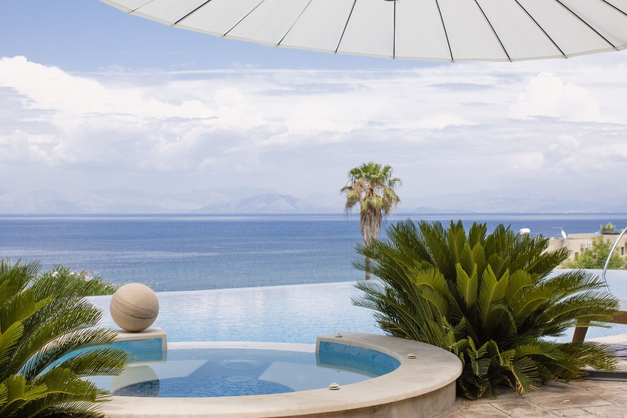A pool area with a view of the sea, palm trees, and a mountain in the background.