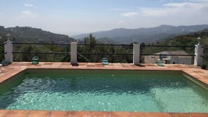 Una piscina rectangular con agua cristalina, rodeada por un suelo de baldosas y una barandilla metálica. Al fondo, se ven colinas y un cielo despejado.