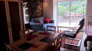 Ein Essbereich mit Holztisch, Stühlen und einer Steinwand. Eine Couch mit roten Kissen und Blick ins Freie durch eine Glastür.