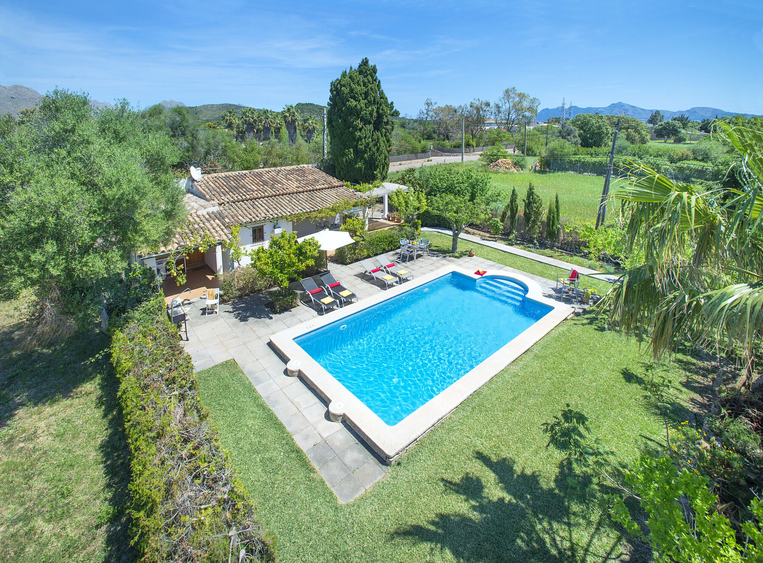 A large rectangular swimming pool with a tiled deck surrounding it, a covered patio area with outdoor furniture, and lush greenery in the background.