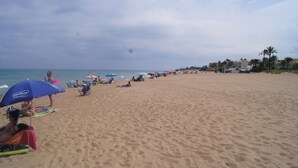 Een strand met mensen die ontspannen onder parasols, een zandstrand en gebouwen op de achtergrond.