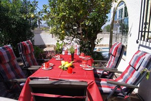 Uma área de refeições ao ar livre com toalha de mesa vermelha, cadeiras listradas e uma mesa posta com pratos, copos e um vaso de flores.