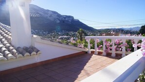 Een terras met uitzicht op een bergachtig landschap, witte balustrades en roze bloemen.