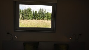 A bedroom with a window overlooking a grassy field and trees. There are two bedside lamps and a bed with pillows.