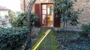 Een stenen gebouw met een houten deur en luiken, een smal pad naar de ingang, en omliggend groen.