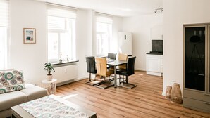 Un salon moderne comprenant un canapé, une table à manger avec des chaises, une petite table d’appoint avec une plante, et un coin cuisine avec des placards et un réfrigérateur.