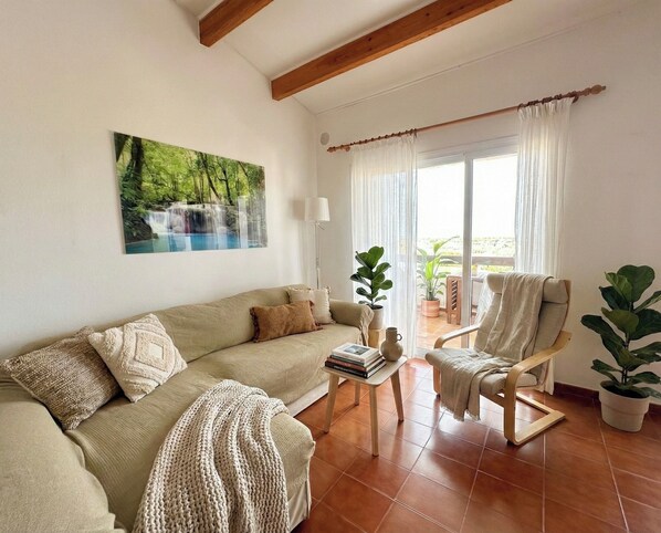 A living room with a sofa, armchair, and a painting of a waterfall. There is a table with books and a lamp. - Cozy apartment in Costa Arenal with sea views (Es Mercadal)