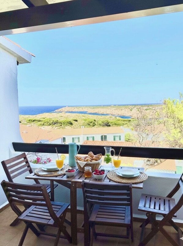 A balcony with a table set for breakfast, overlooking a coastal landscape.