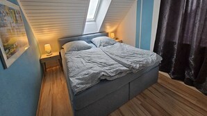 A bedroom with a sloped ceiling, a skylight, a bed with a grey duvet, a nightstand with a lamp, and a dark curtain. - Family apartment with sea view (Fehmarn)