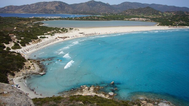 Un paisaje costero con playa, aguas azules cristalinas y montañas al fondo.