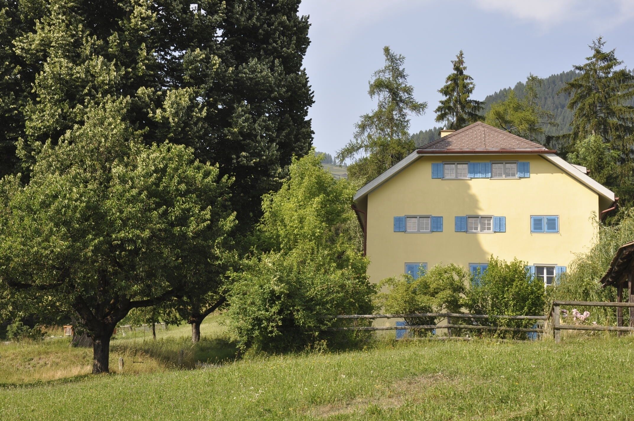 Ein gelbes Gebäude mit blauen Fensterläden, umgeben von Bäumen und einem Holzzaun.