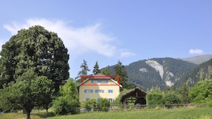 Un bâtiment à deux étages, doté d’une façade jaune et de volets bleus, entouré d’arbres et de montagnes.