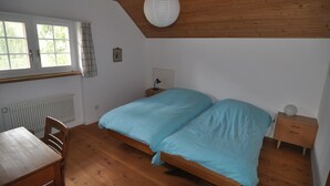 Une chambre avec un plafond en bois, une fenêtre avec des rideaux, un lit recouvert d’une couverture bleue, une table de chevet avec une lampe et un radiateur.