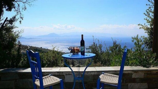 Ein Tisch mit einer Flasche und einer Schüssel, zwei blaue Stühle und Blick auf das Meer und die Berge.