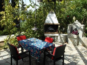An outdoor patio with a table covered in a floral tablecloth, surrounded by wicker chairs with red cushions. - detached holiday house, calmly located in an olive grove (Thessalien)