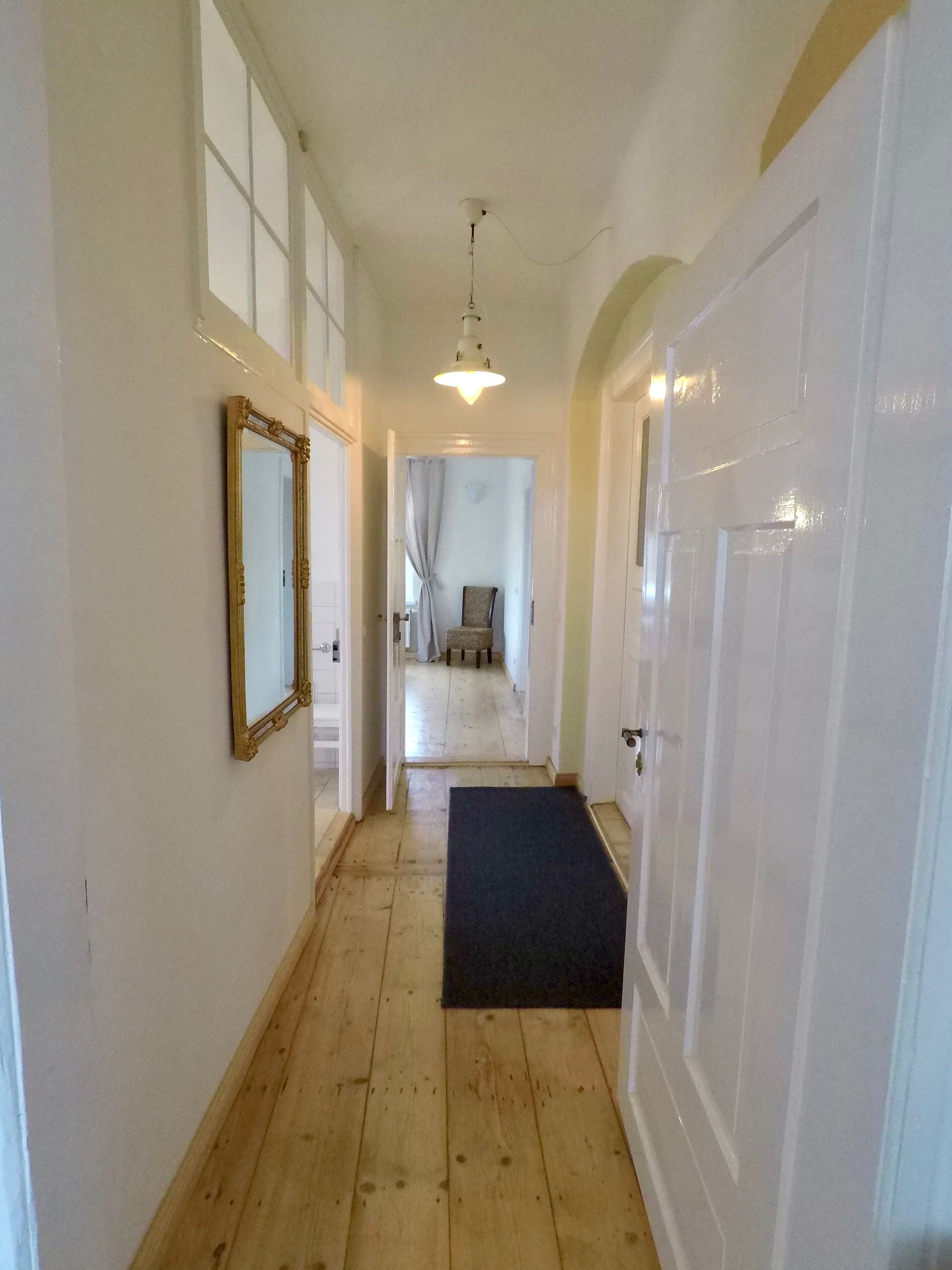 A hallway with a wooden floor, a black doormat, a mirror on the left wall, and a door on the right.