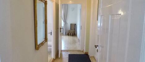 A hallway with a wooden floor, a black doormat, a mirror on the left wall, and a door on the right.