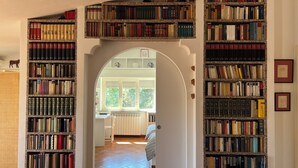 Una biblioteca con numerosas estanterías llenas de libros, que conduce a una habitación con ventanal y radiador.