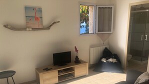 Ein Wohnzimmer mit Fernseher auf einem Holzboden, einem kleinen Tisch, einer Couch und einem Fenster mit Blick auf Bäume und ein Gebäude.