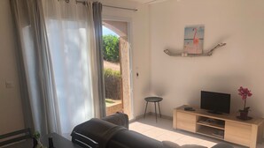 Ein Wohnzimmer mit einer Couch, einem Fernseher auf einem Holzboden, einem kleinen Tisch mit Flaschen und Blick durch eine Glastür.