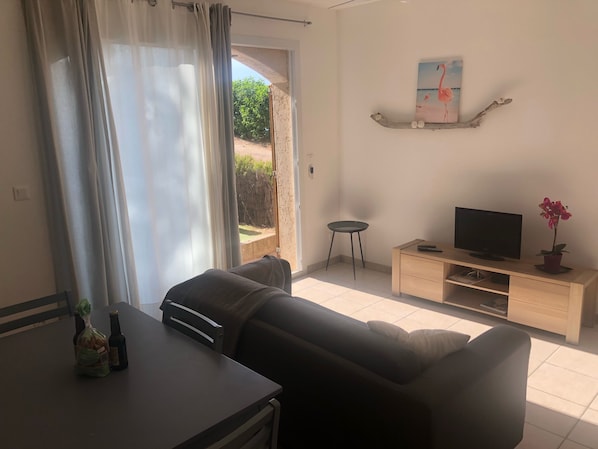 A living room with a sofa, a TV on a wooden stand, a small table with bottles, and a view through a glass door. - RENTAL CORSICA, T2, Haute-Corse, BALAGNE, CORBARA, 2 pers, Calm and rest assured (Corbara)