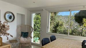 A dining area with a table covered in a floral tablecloth, a wicker chair with a cushion, and a large window with a view of greenery and hills. - Beautiful sea view - Spacious and comfortable - Walking distance to the beach (Le Lavandou)