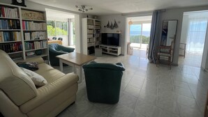 A living room with a sofa, a green armchair, a coffee table, and a TV. There are bookshelves filled with books and a ceiling fan. - Beautiful sea view - Spacious and comfortable - Walking distance to the beach (Le Lavandou)