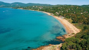 An aerial view of a coastal area with a sandy beach, rocky shoreline, and a backdrop of green hills with scattered buildings. - Provençal villa with sea views & Mediterranean garden, beaches, Wi-Fi, A/C (La Croix-Valmer)