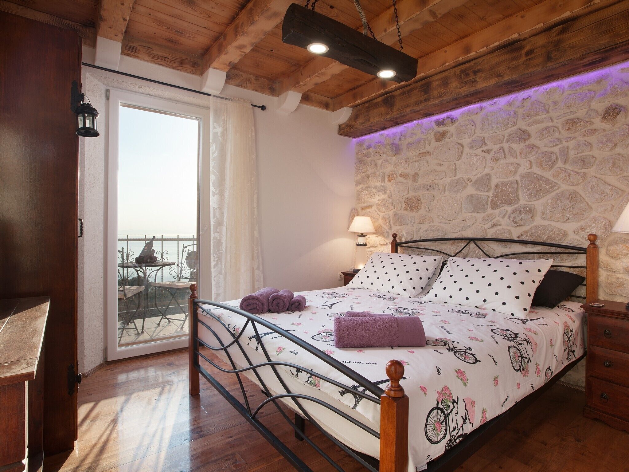 Une chambre avec un plafond en bois, un mur en pierre, un lit recouvert d’une couvre-lit à motifs de vélos, et un balcon avec une table et des chaises.