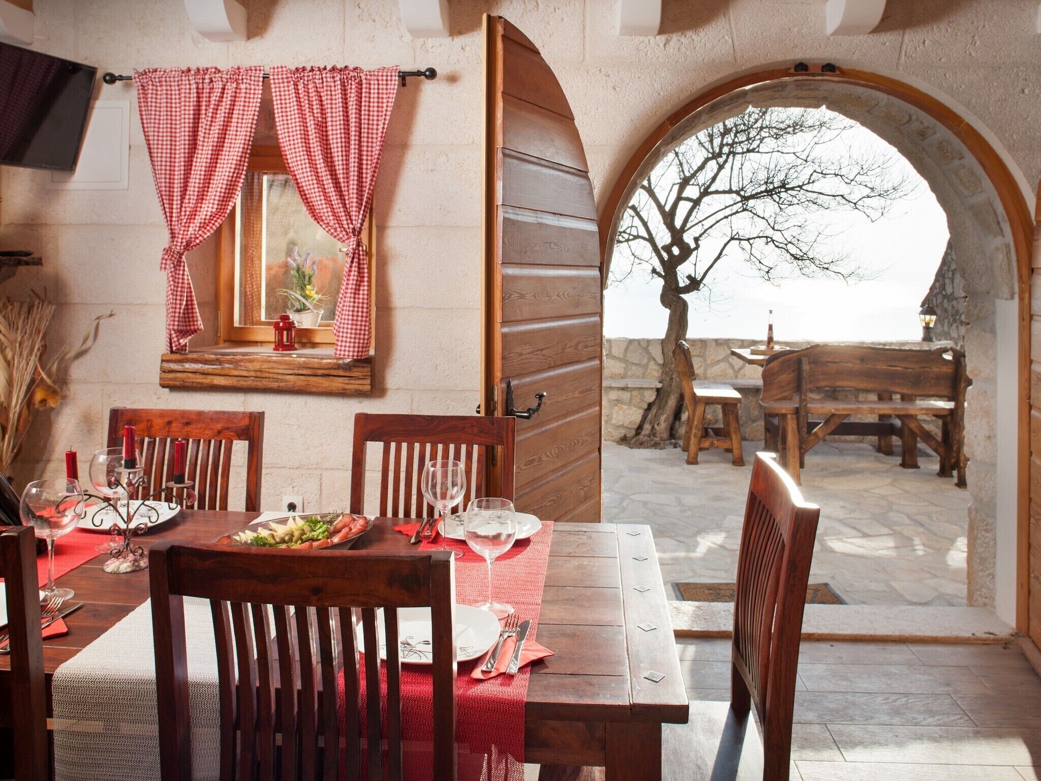 Un coin repas avec une table en bois dressée pour deux personnes, un miroir au-dessus de la table et, à travers une porte en arc de cercle, une vue sur un espace extérieur aménagé pour s’asseoir.
