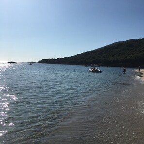 Une plage à l’eau claire, un bateau et un paysage vallonné en arrière-plan.